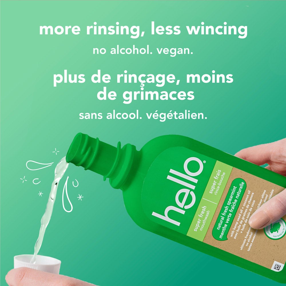 A hand pours hello brand green super fresh mouthwash into the mouthwash bottle cap with a light green background. The bottle label is clear. White text reads, "more rinsing, less wincing – no alcohol. vegan." with French translation below.