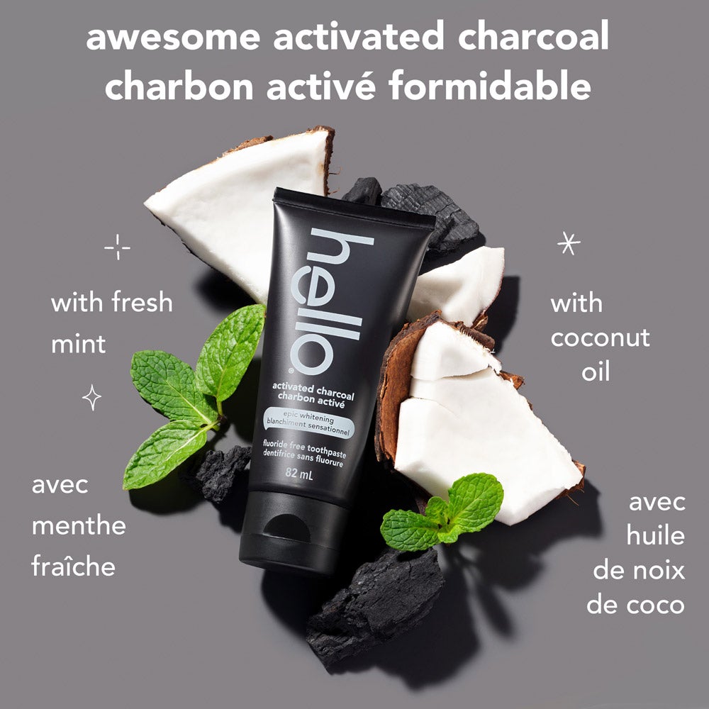 A black tube of hello epic whitening fluoride free charcoal toothpaste is centered, surrounded by pieces of coconut, black charcoal chunks, and fresh mint leaves. The background is light grey. Text above the product reads &quot;awesome activated charcoal.&quot; To the left, it says &quot;with fresh mint,&quot; and to the right, &quot;with coconut oil.&quot;