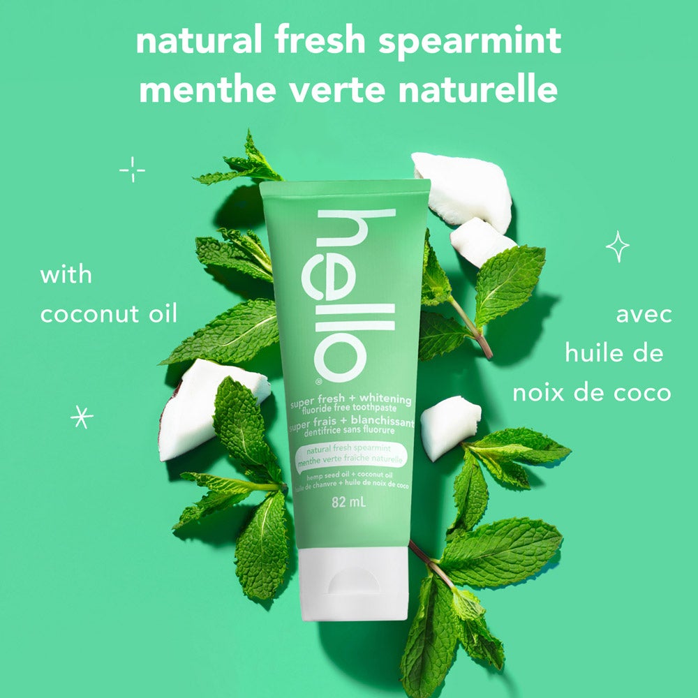 A green tube of hello super fresh + whitening fluoride free toothpaste stands at the center on a solid green background, surrounded by fresh mint sprigs and coconut pieces. Above the product, the text reads: "natural fresh spearmint." To the left, it says "with coconut oil."