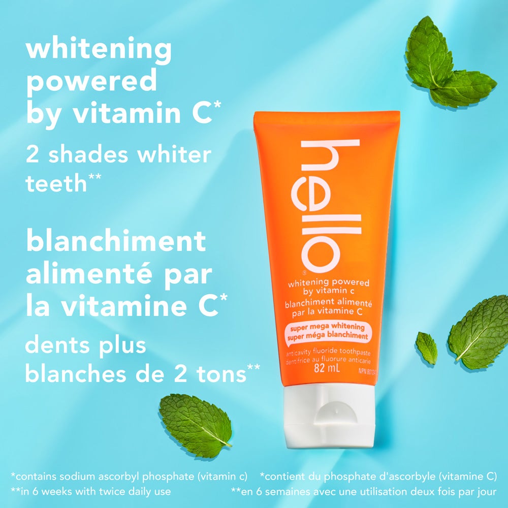An orange tube of hello super mega whitening toothpaste stands upright on a light blue background scattered with fresh mint leaves. Text highlights "whitening powered by vitamin C" and "2 shades whiter teeth" in English and French, with additional small print disclaimers at the bottom.